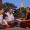 ミャンマーの伝統的な寺院で、ビルマ暦の曜日ごとの命名ルールについて真剣な表情で話を聞く日本人家族。僧侶が伝統的なカレンダーを指差している。