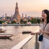 夕暮れ時の美しいチャオプラヤー川とワット・アルンを背景に、安全対策を意識した斜め掛けバッグを身につけ、笑顔で景色を楽しむ日本人の女性旅行者。