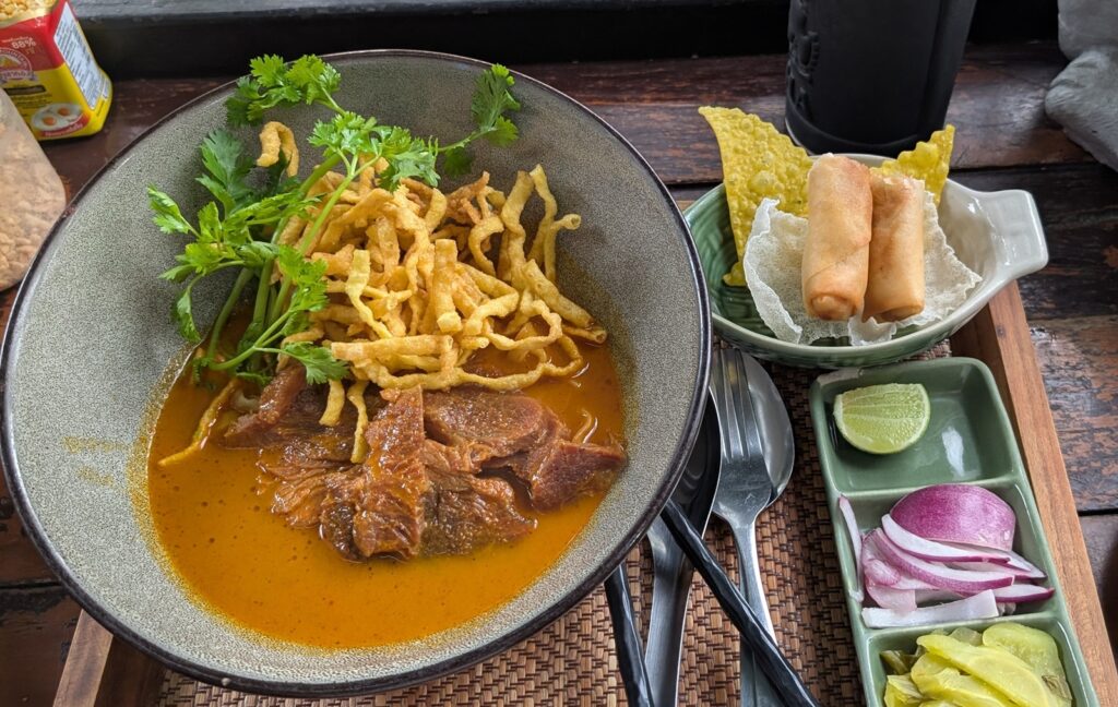 タイ・チェンマイ名物のカオソーイ。カレー風味のスープ麺に揚げ麺をのせ、ライムや紫タマネギなどを添えた料理