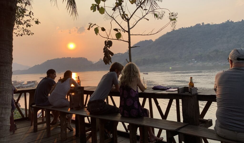 メコン川沿いのテラス席から山の向こうに沈む夕日を眺める旅行者