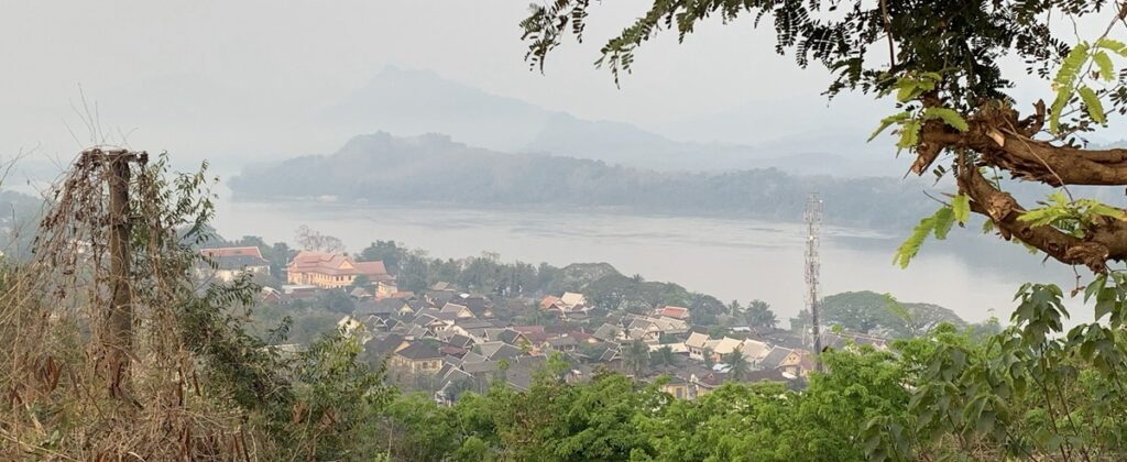 ルアンパバーンの高台から市街地と川、周囲の山々を見渡した風景。手前には伝統的な屋根の建物が密集し、奥には霞に包まれた山並みが続く。