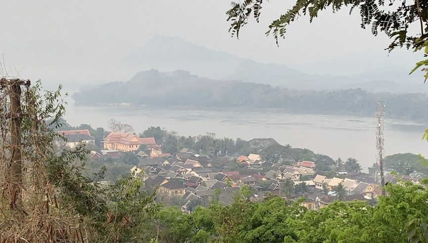 ルアンパバーンの高台から市街地と川、周囲の山々を見渡した風景。手前には伝統的な屋根の建物が密集し、奥には霞に包まれた山並みが続く。
