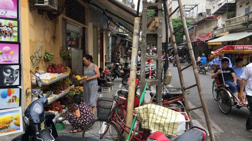 ハノイ旧市街の細い通りに果物の露店や自転車、バイク、シクロが並ぶ街並み