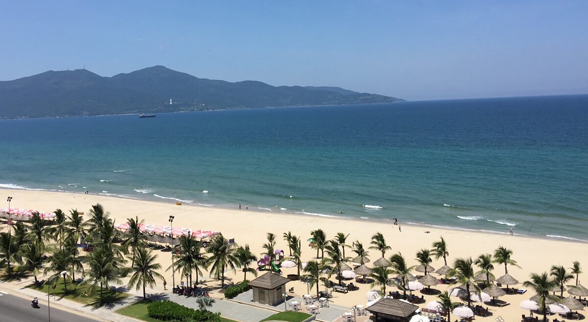 高台から見る、青く広がる海と白い砂浜、ヤシの木が並ぶリゾートエリア。遠くには山並みが見え、開放感のある景色が広がっている。