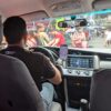 運転席のドライバーやナビゲーション用スマートフォンが写っており、フロントガラス越しには多くの歩行者や露店でにぎわう街並みが見える