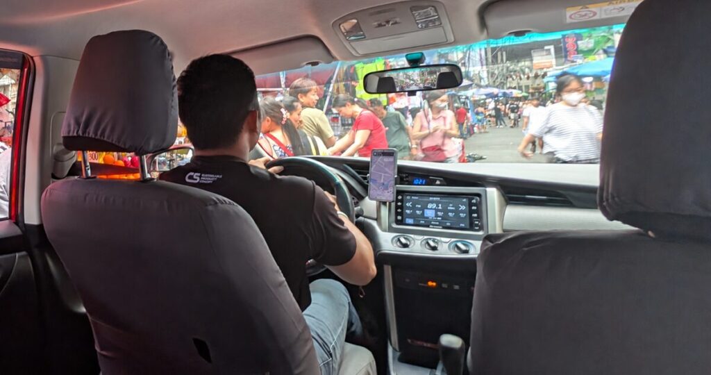 運転席のドライバーやナビゲーション用スマートフォンが写っており、フロントガラス越しには多くの歩行者や露店でにぎわう街並みが見える