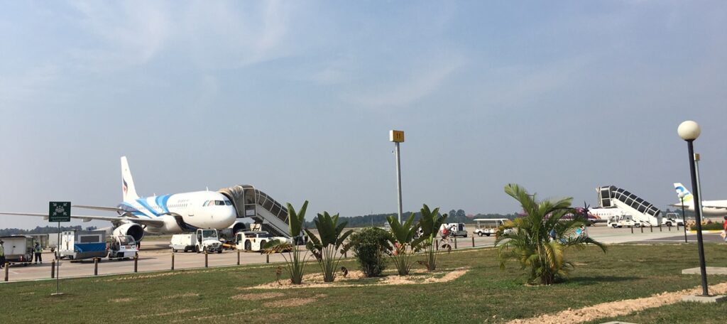 旅客機が駐機し、搭乗用タラップや空港作業車が見える。手前には南国らしいヤシの木や緑地が広がっている。