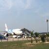 旅客機が駐機し、搭乗用タラップや空港作業車が見える。手前には南国らしいヤシの木や緑地が広がっている。